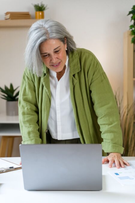 Confident senior woman smiling while working on her laptop and reviewing paperwork at her desk